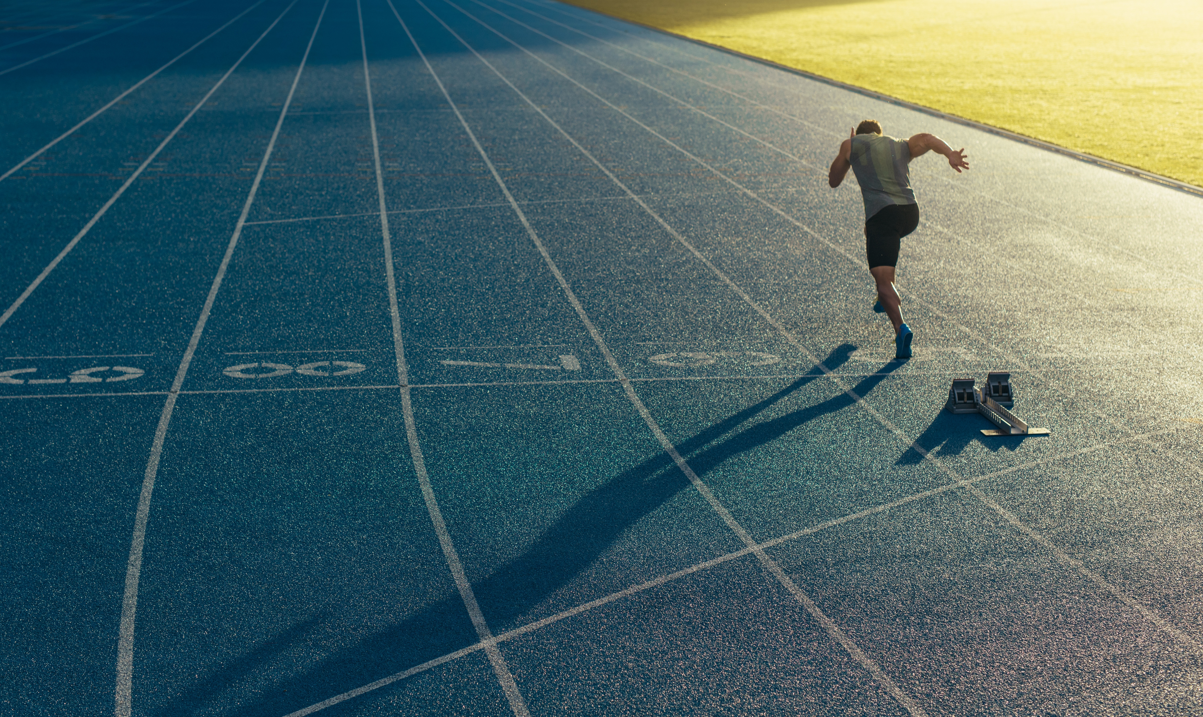 An athlete training with starting blocks at a bright blue athletics track at dusk.