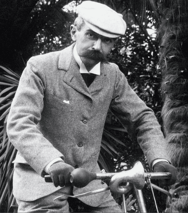 A black and white photograph of Pierre De Coubertin, cycling while wearing a suit.